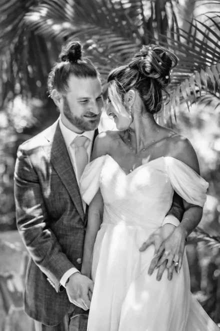 Black and white wedding photography in Puerto Vallarta. Close-up portrait of a bride and groom smiling intimately at each other, surrounded by tropical palm leaves
