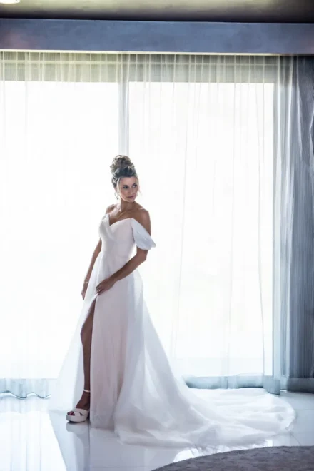 Bridal photography in Puerto Vallarta. Bride getting ready session before the wedding, wearing a white off-the-shoulder dress in front of a bright window