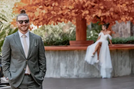 Destination wedding photography in Puerto Vallarta. A joyful groom with sunglasses and a hair bun smiles in the foreground, with his bride sitting gracefully in the background under a vivid orange tree