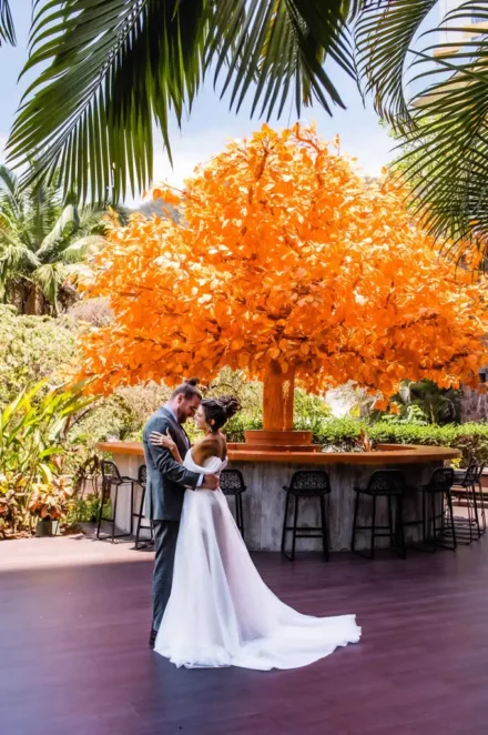 Luxury wedding photography in Puerto Vallarta. Newlywed couple embracing on an outdoor terrace framed by palm leaves, featuring a stunning bright orange decorative tree and a modern circular bar in the background