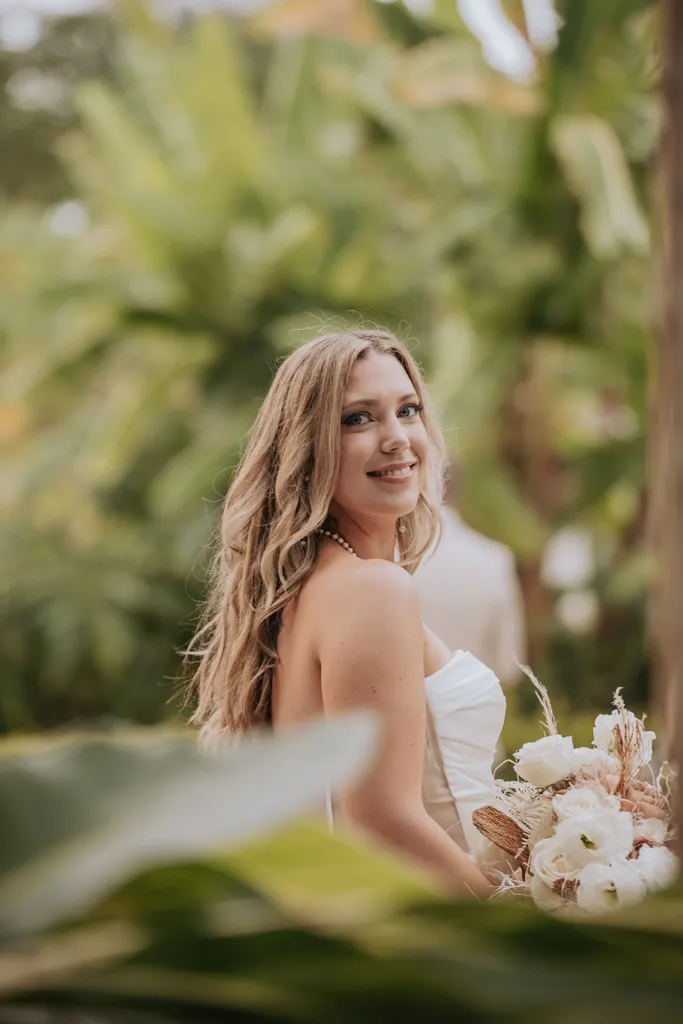 A beautiful blonde bride smiling over her shoulder while holding a white roses bouquet in a lush tropical garden setting in Puerto Vallarta
