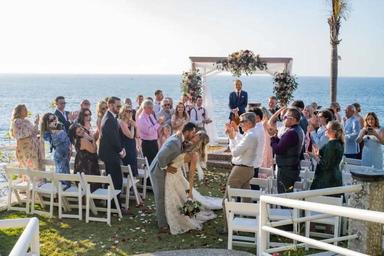 A groom dips his bride for a romantic first kiss during an outdoor oceanfront wedding ceremony in Puerto Vallarta, as wedding guests stand and applaud in an open-air venue by the sea