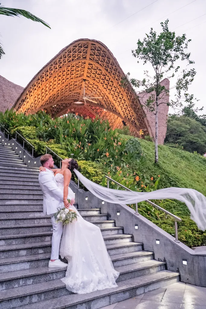 A joyful groom in a white suit holds his bride on a grand stone staircase during a wedding in Puerto Vallarta. The bride's long tulle veil caught in the wind blows elegantly to the side, with a massive, beautifully lit tropical wooden palapa pavilion structure in the background