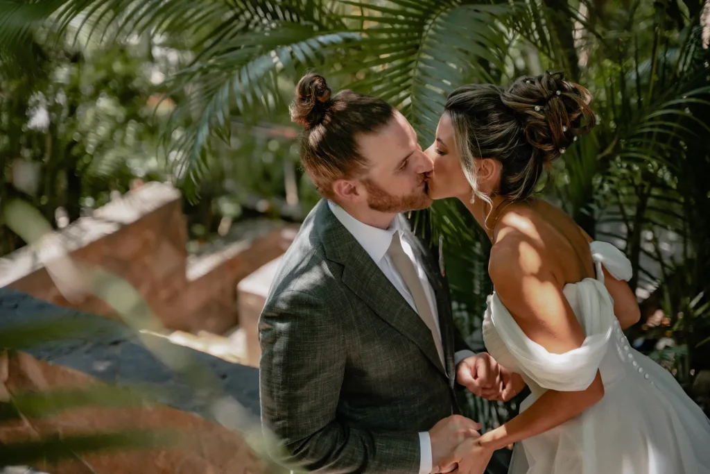 Close-up portrait of a bride and groom holding hands and kissing during their destination wedding in Puerto Vallarta. The groom has a hair bun and a red beard, wearing a textured grey suit with a beige tie. The bride features an elegant updo hairstyle with pearls, wearing an off-the-shoulder white wedding dress. Sunbeams and shadows filter through lush green palm leaves in the background