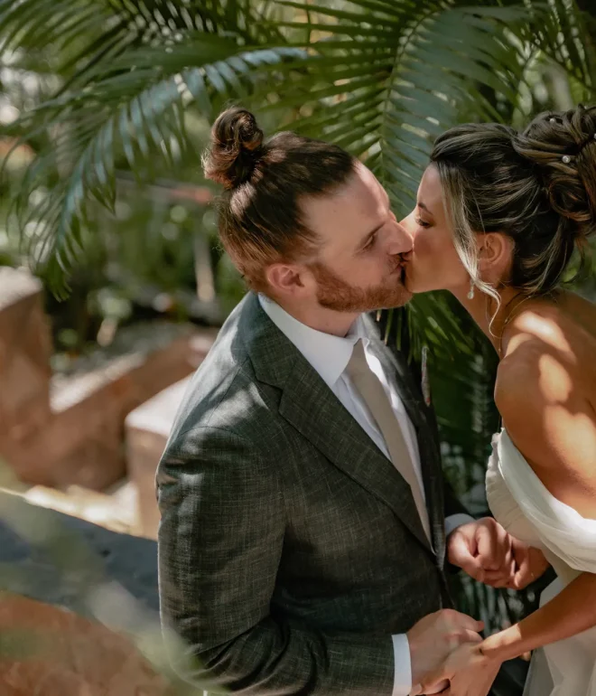 Close-up portrait of a bride and groom holding hands and kissing during their destination wedding in Puerto Vallarta. The groom has a hair bun and a red beard, wearing a textured grey suit with a beige tie. The bride features an elegant updo hairstyle with pearls, wearing an off-the-shoulder white wedding dress. Sunbeams and shadows filter through lush green palm leaves in the background