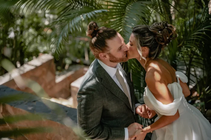 Close-up portrait of a bride and groom holding hands and kissing during their destination wedding in Puerto Vallarta. The groom has a hair bun and a red beard, wearing a textured grey suit with a beige tie. The bride features an elegant updo hairstyle with pearls, wearing an off-the-shoulder white wedding dress. Sunbeams and shadows filter through lush green palm leaves in the background