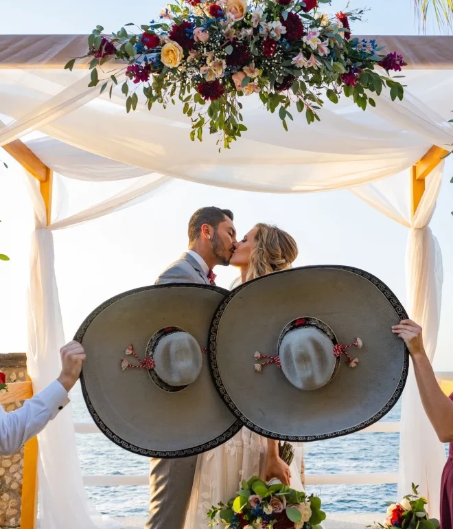 A bride and groom sharing a romantic kiss under a floral wedding altar next to the ocean in Puerto Vallarta, playfully framed by bridesmaids and groomsmen holding large traditional Mexican mariachi hats