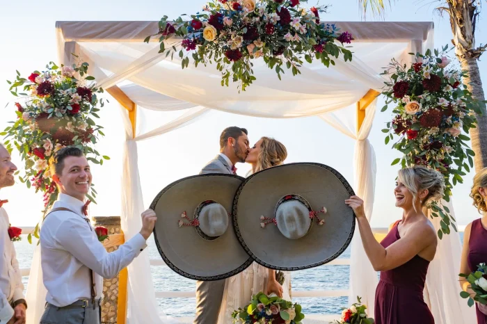 A bride and groom sharing a romantic kiss under a floral wedding altar next to the ocean in Puerto Vallarta, playfully framed by bridesmaids and groomsmen holding large traditional Mexican mariachi hats