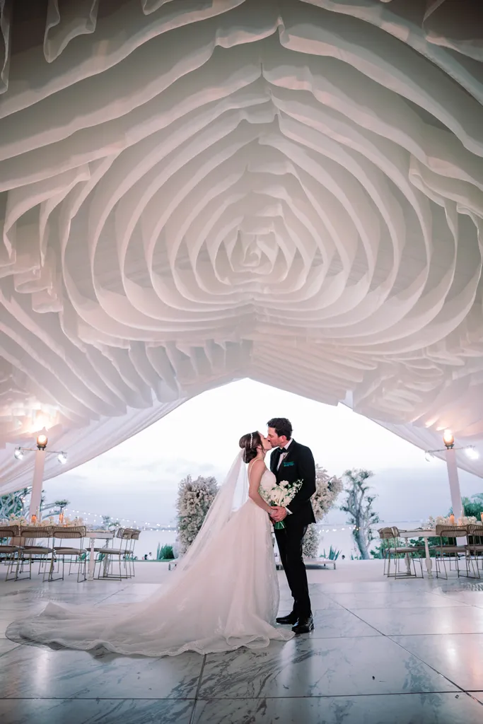 A bride and groom sharing a romantic kiss on a marble dance floor during their luxury wedding reception in Puerto Vallarta. They stand under a stunning, massive white fabric ceiling installation draped intricately in the shape of a blooming rose, with a coastal twilight background