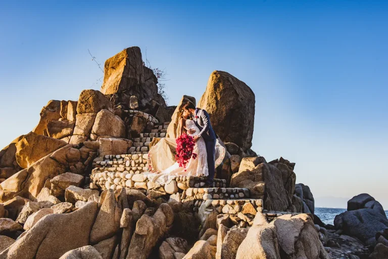 A bride and groom kissing on stone steps carved into massive oceanfront rocks during a wedding photo shoot under a clear blue sky in Puerto Vallarta