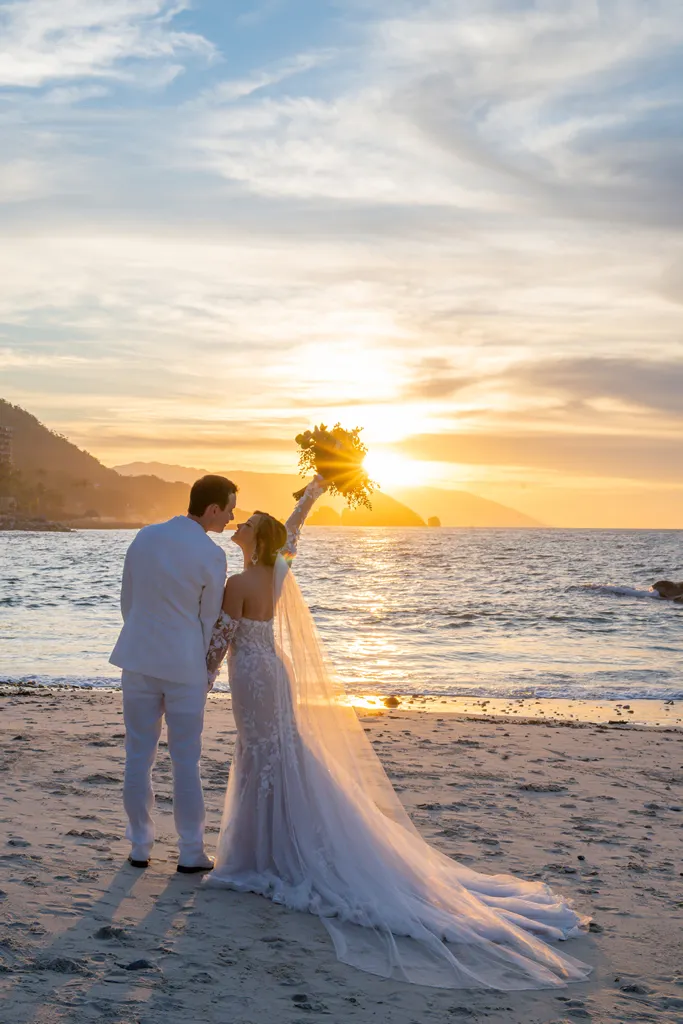 Newlyweds standing on a sandy beach during a golden hour sunset photo shoot in Puerto Vallarta, with the bride holding her bouquet up