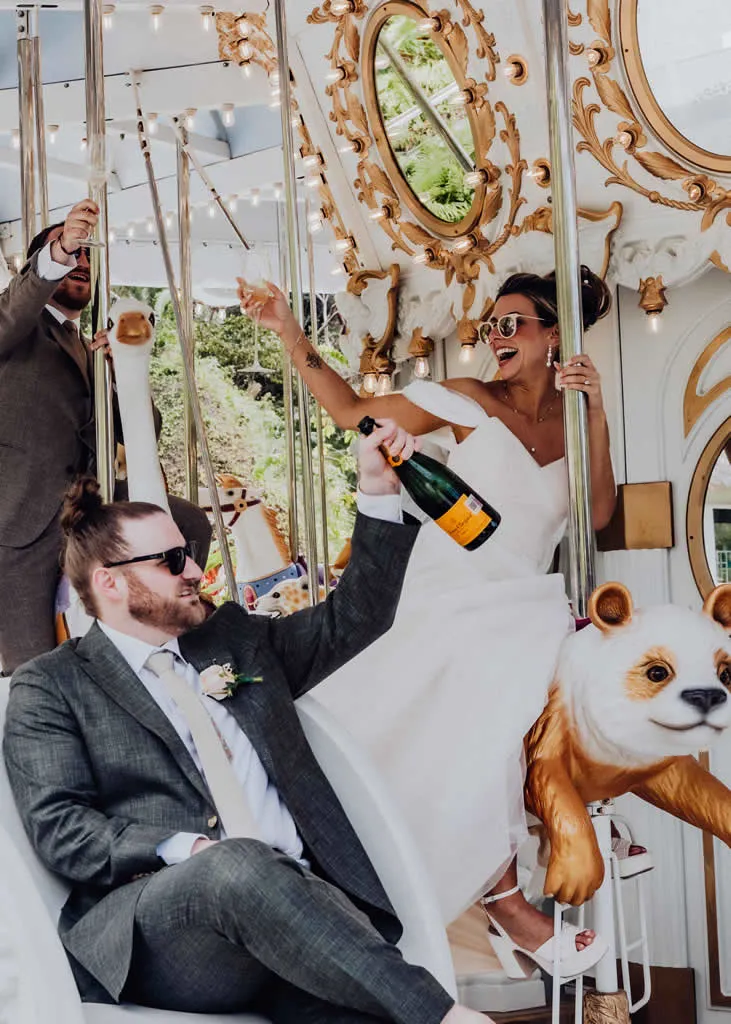 A joyful bride and groom celebrating with champagne glasses and wearing sunglasses on a vintage carousel at TierraLuna Gardens in Puerto Vallarta
