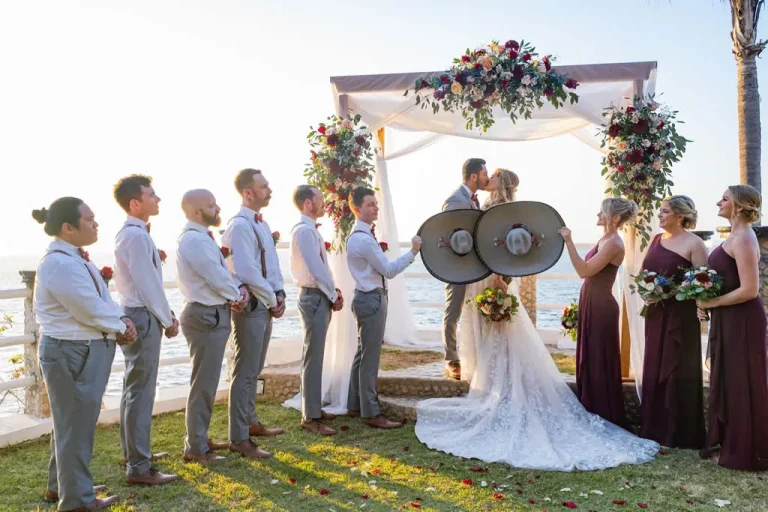 A wide shot of a wedding party standing on a grassy lawn next to the ocean in Puerto Vallarta. The bride and groom kiss under a white draped floral altar, while a groomsman and a bridesmaid playfully hold up two large traditional Mexican mariachi hats