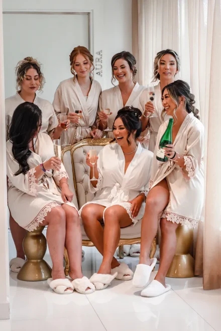 Bride seated in a beige silk robe surrounded by her bridesmaids holding champagne glasses in a joyful wedding toast