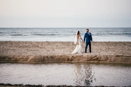A serene wide-angle shot by the ocean shore. Holding hands and gazing affectionately at each other, the newlyweds take a quiet stroll along the beach, surrounded by the gentle rhythm of the waves in Puerto Vallarta