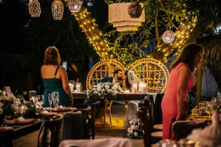 A magical evening filled with warmth and romance. Seated under the soft glow of fairy lights and woven lanterns, the newlyweds share a sweet kiss at their sweetheart table, surrounded by the joy and laughter of their guests in Puerto Vallarta