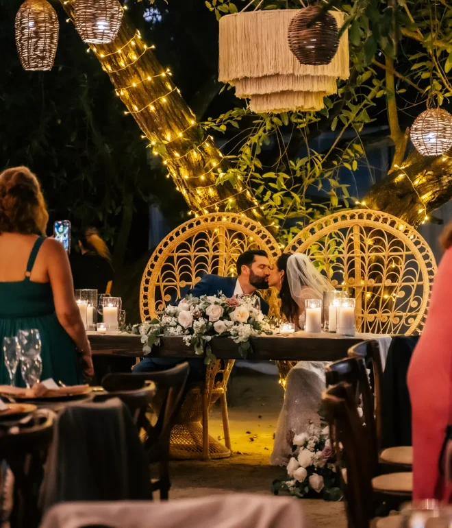 A magical evening filled with warmth and romance. Seated under the soft glow of fairy lights and woven lanterns, the newlyweds share a sweet kiss at their sweetheart table, surrounded by the joy and laughter of their guests in Puerto Vallarta