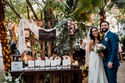 A charming personal touch surrounded by tropical beauty. Standing beside their rustic welcome setup—complete with seating cards, romantic floral accents, and glowing candles—the happy couple welcomes their guests to an unforgettable celebration in Puerto Vallarta