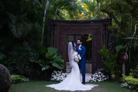 Framed by nature and timeless craftsmanship. Standing beneath a grand carved wooden doorway surrounded by lush palm foliage, the newlyweds exchange a loving look during their celebration in Puerto Vallarta