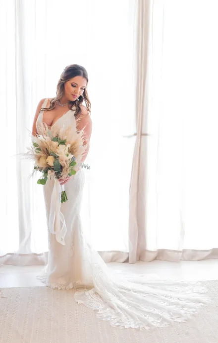 A glowing portrait of serene beauty and effortless tropical romance. Holding a delicate bohemian bouquet, the bride reflects in quiet anticipation, framed by soft beachside window light before stepping toward her forever in Puerto Vallarta