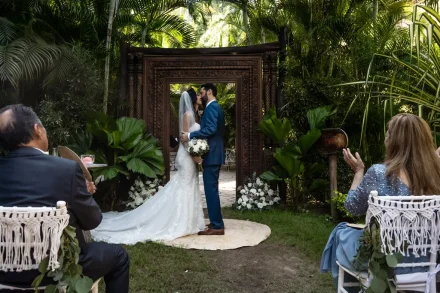 Sealed with a kiss in a lush tropical paradise. Surrounded by vibrant greenery and loved ones, the newlyweds celebrate their first kiss as husband and wife under an ornate wooden arch in Puerto Vallarta
