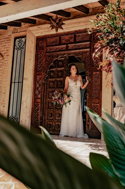 A peaceful moment of sweet anticipation before taking the biggest step. Framed by grand rustic architecture and lush greenery, the bride stands gracefully in the doorway, holding her bouquet in an unforgettable atmosphere in Puerto Vallarta