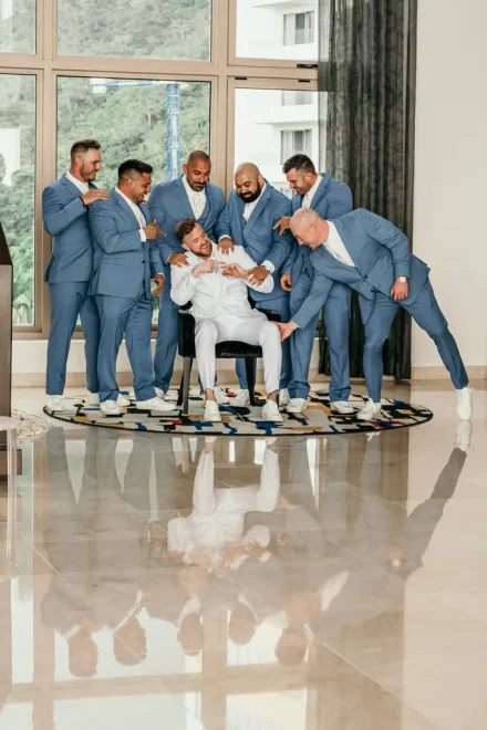 Groom in a white suit sitting as groomsmen in light blue suits point playfully at his wedding ring