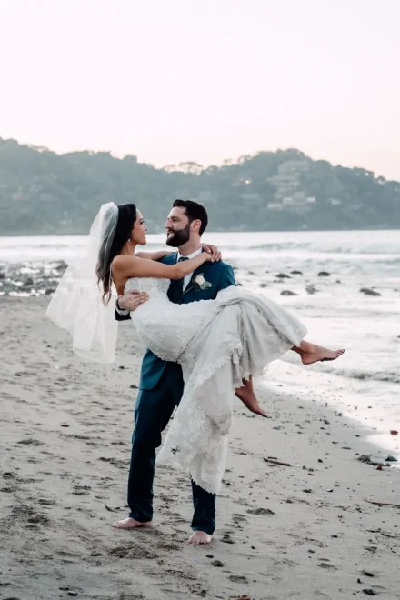 A romantic escape to the shore! Walking barefoot along the sand as the waves roll in, the groom carries his smiling bride in his arms for a timeless beach portrait in Puerto Vallarta