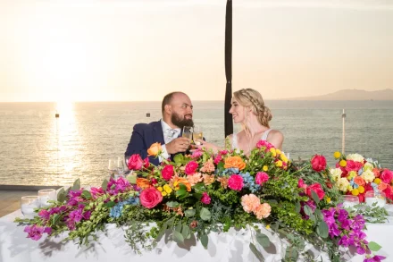 A toast to forever set against a glowing horizon. Seated together behind a cascade of vibrant floral arrangements, the newlyweds share a gentle gaze and clink champagne glasses as the sun dips over the Pacific in Puerto Vallarta