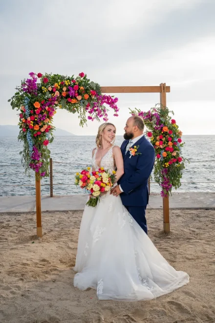 A tender look amidst vibrant tropical beauty. Standing hand in hand beneath a lush wooden arch overflowing with colorful blooms, the newly married couple shares an intimate gaze against the sparkling Pacific ocean in Puerto Vallarta