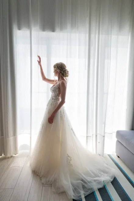 Ethereal grace in full bloom. Standing fully dressed in her flowing lace gown, gently touching sheer curtains as bright light fills the room, this captivating bridal portrait embodies grace, beauty, and quiet romance in Puerto Vallarta