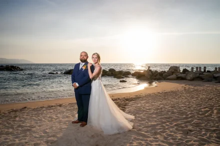 Bathed in the warm glow of the setting sun. Resting her hand gently upon his shoulder, the bride and groom pose together along the tranquil shoreline as golden light reflects off the gentle ocean waves in Puerto Vallarta