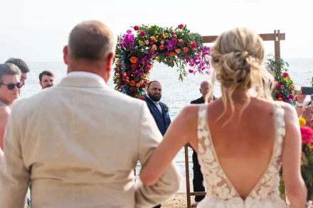 A moment filled with raw, overwhelming emotion. Seen from behind as she walks down the aisle, the groom looks back at his bride with tears in his eyes beneath a vibrant tropical floral arch—capturing the pure heart of a Puerto Vallarta wedding ceremony