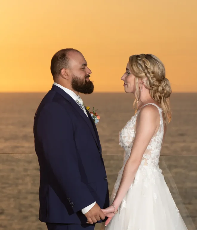 Enveloped in the golden warm glow of twilight. Standing hand-in-hand on a high-rise oceanfront terrace, the newly married couple shares a quiet, romantic moment as the sun dips below the horizon in Puerto Vallarta