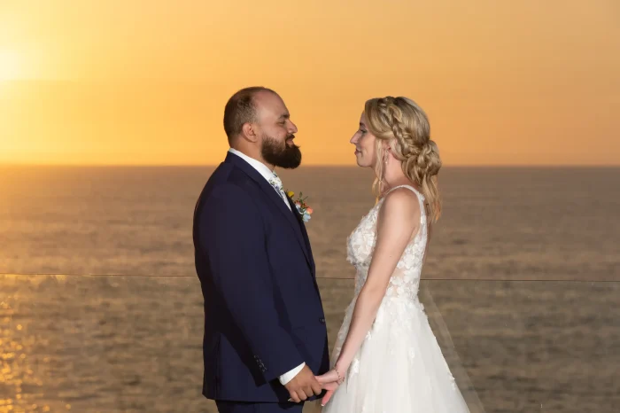 Enveloped in the golden warm glow of twilight. Standing hand-in-hand on a high-rise oceanfront terrace, the newly married couple shares a quiet, romantic moment as the sun dips below the horizon in Puerto Vallarta