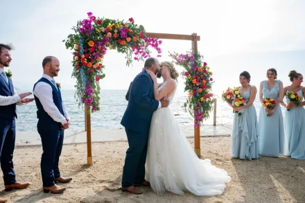 Sealed with a passionate first kiss. Surrounded by the gentle sound of ocean waves and brilliant floral blooms, the newly married couple shares a tender moment under the beachside arch in Puerto Vallarta